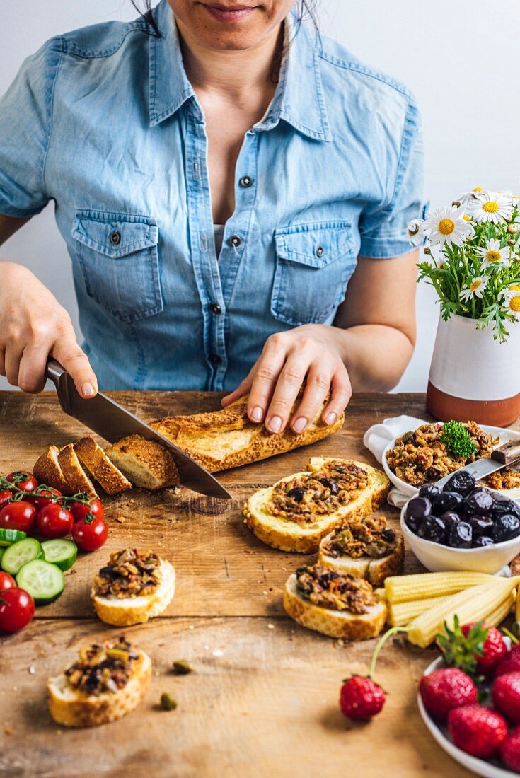 A woman slicing French baguette, olive tapenade on bread slices, olives, pickled baby corns, strawberries