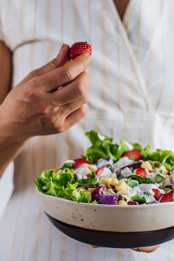 A woman holding a bowl of fresh strawberry pasta salad drizzled with yogurt in one hand and grabbing a strawberry with the other