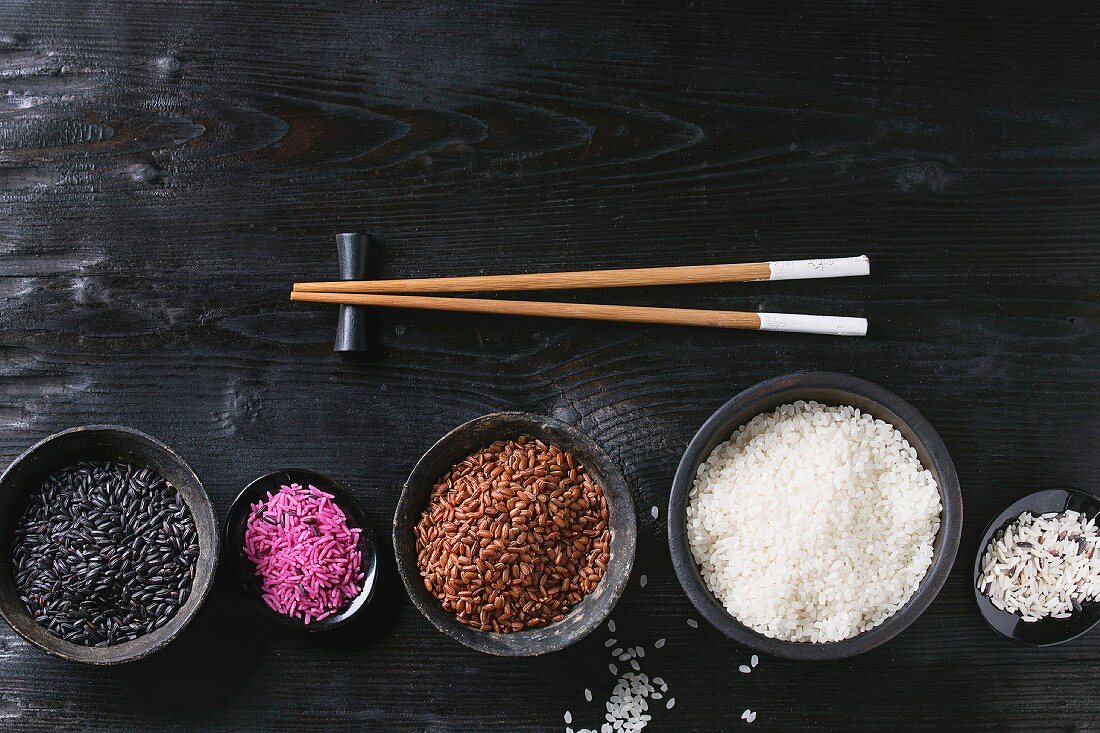 Variety assortment of raw uncooked colorful rice white, black, brown, pink in black bowls over wooden background