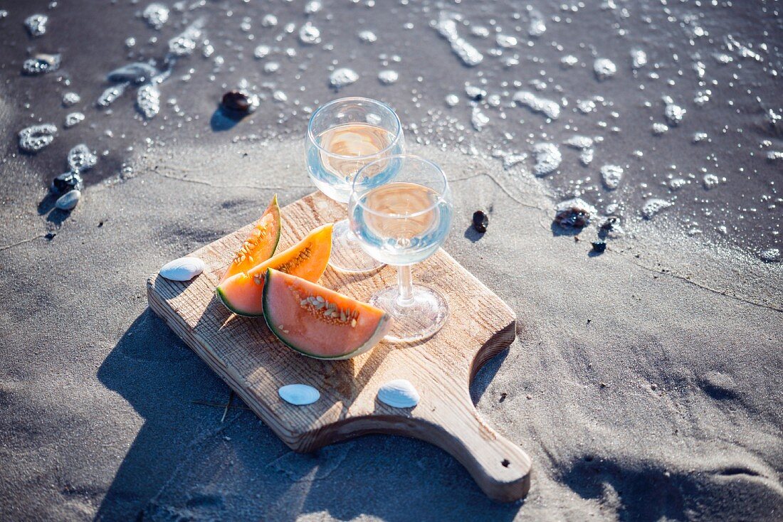 Two glasses of white wine and cantaloupe melon slices on a sandy beach