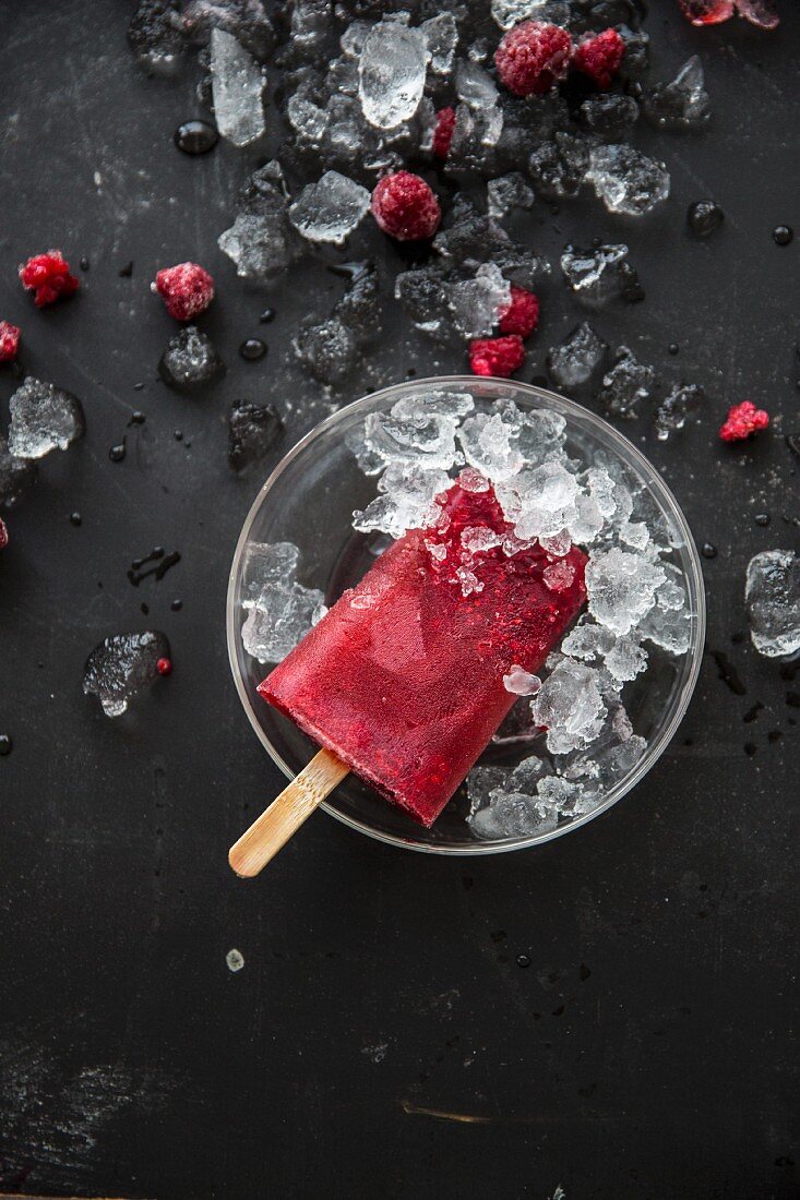 A raspberry ice lolly on a stick in a bowl of crushed ice