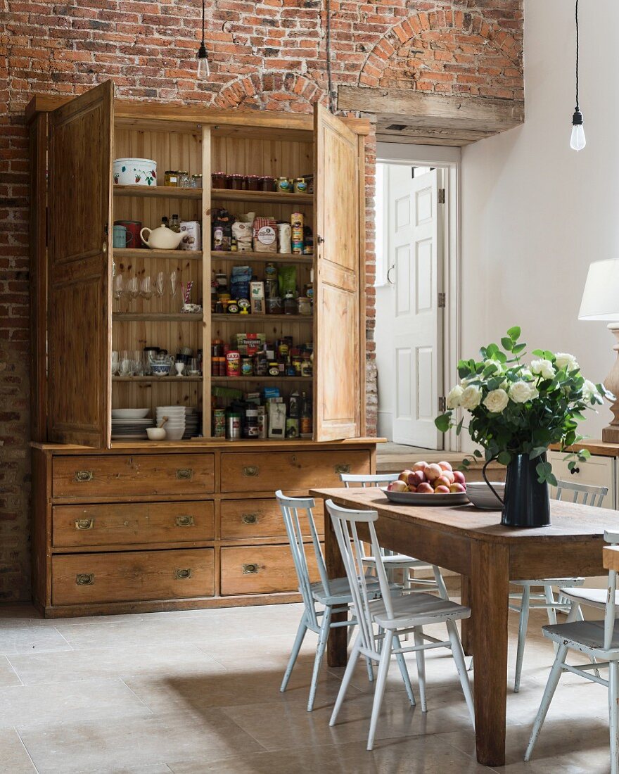 Rustic wooden cupboard with open doors and dining table in spacious kitchen with exposed brick wall