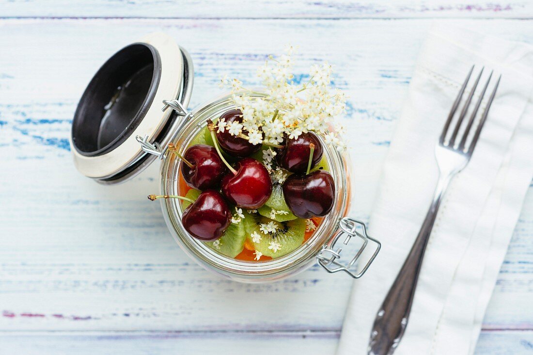 Fruit salad in jar with grapes, mango, banana, cherries, apricot, kiwi, garnished with elder flowers
