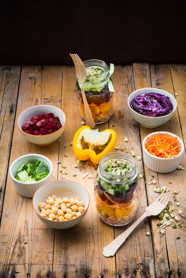 Regenbogen-Salat mit Kichererbsen, Sprossen und verschiedenen Gemüse im Glas, umgeben von Zutaten