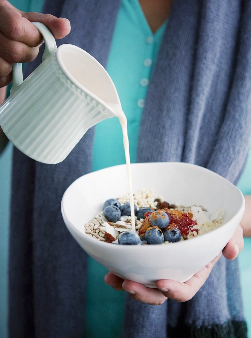A woman pouring an oat drink over gluten-free millet muesli with blueberries