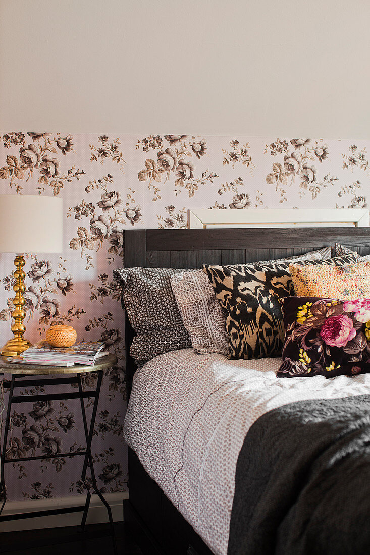 Scatter cushions on double bed and table lamp on bedside table against patterned wallpaper