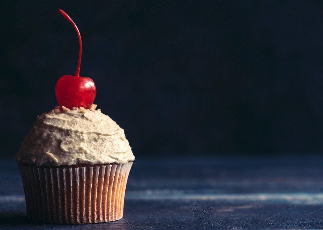 A cupcake with peanut frosting and a cocktail cherry in front of a dark background