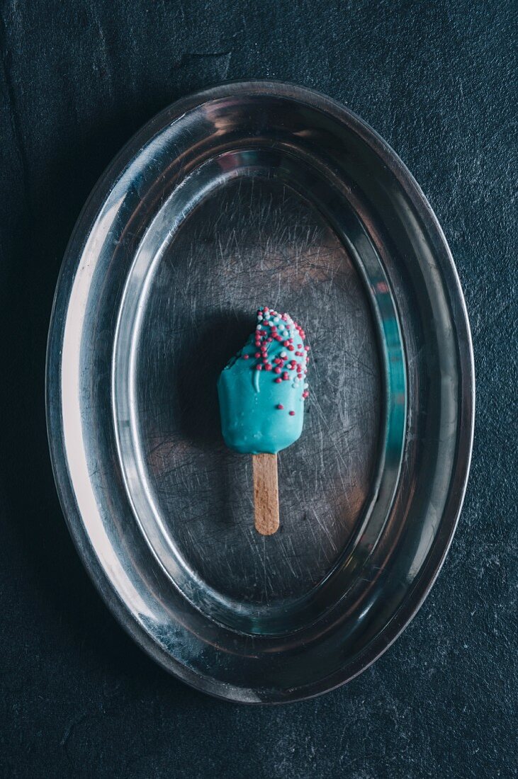 A cake pop in the shape of an ice lolly with brightly coloured icing on a silver tray