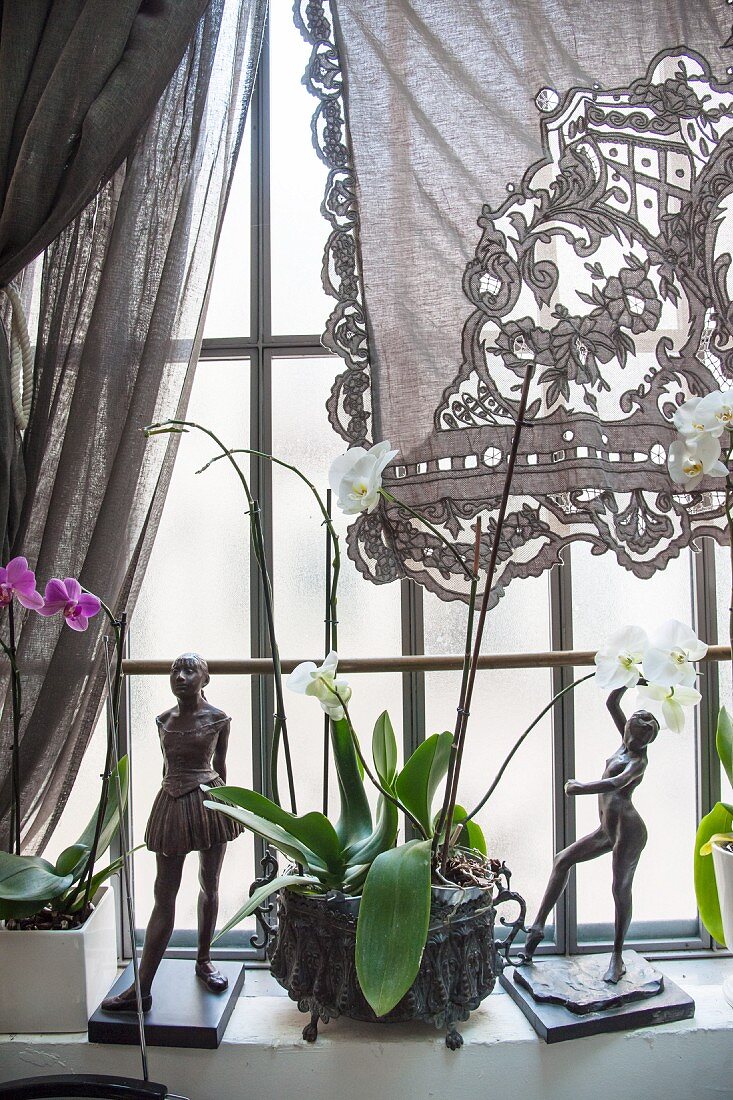 Two sculptures and orchid on windowsill below lace curtain