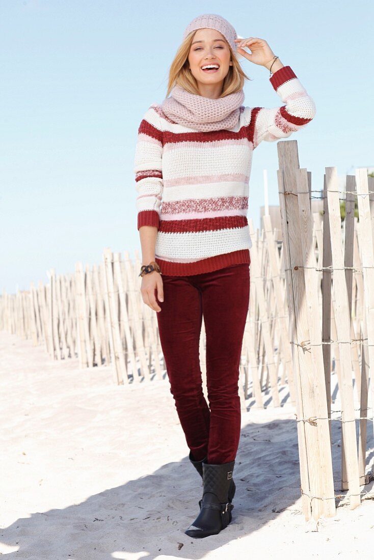 A blonde woman wearing a striped jumper, dark red trousers, a knitted hat and a scarf