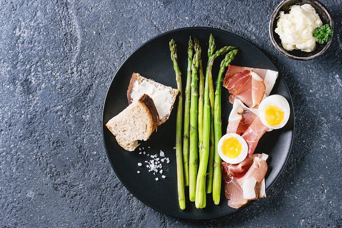 Green asparagus with egg, bread and ham bacon on a black plate (seen from above)