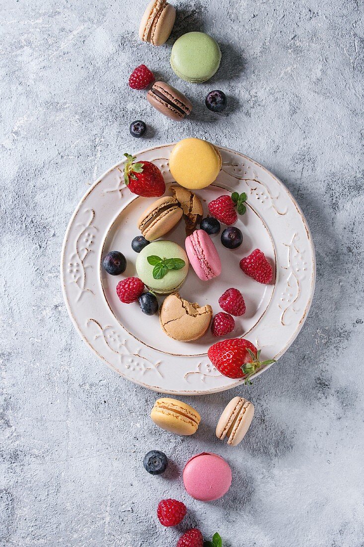 Variety of colorful french sweet dessert macarons with different fillings served on white vintage plate with spring flowers and berries