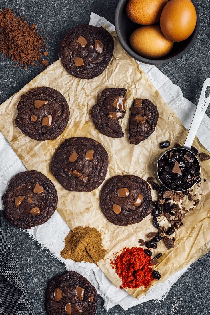 Mexican hot chocolate cookies on a piece of light brown paper view with a cup of chocolate chips, eggs, cayenne pepper, cocoa powder and cinnamon