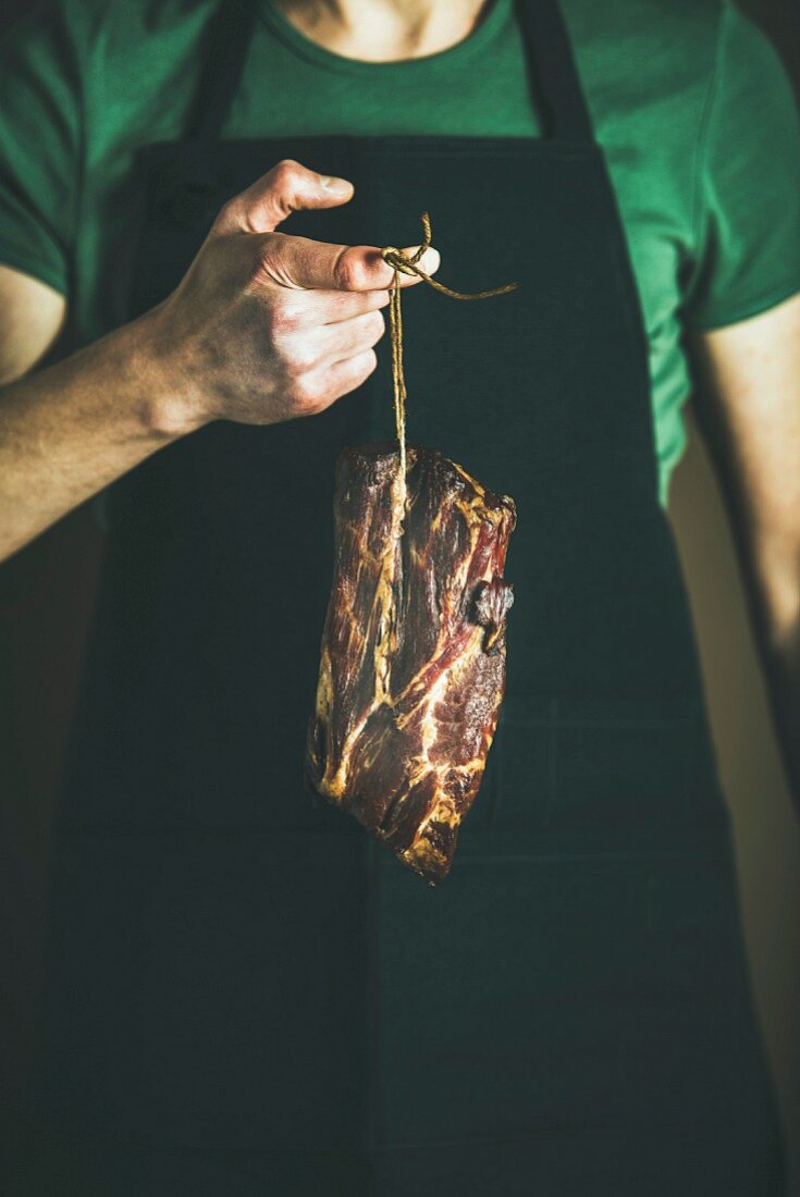 Man in black apron keeping cut of cured pork meat in his hand at local farmers market