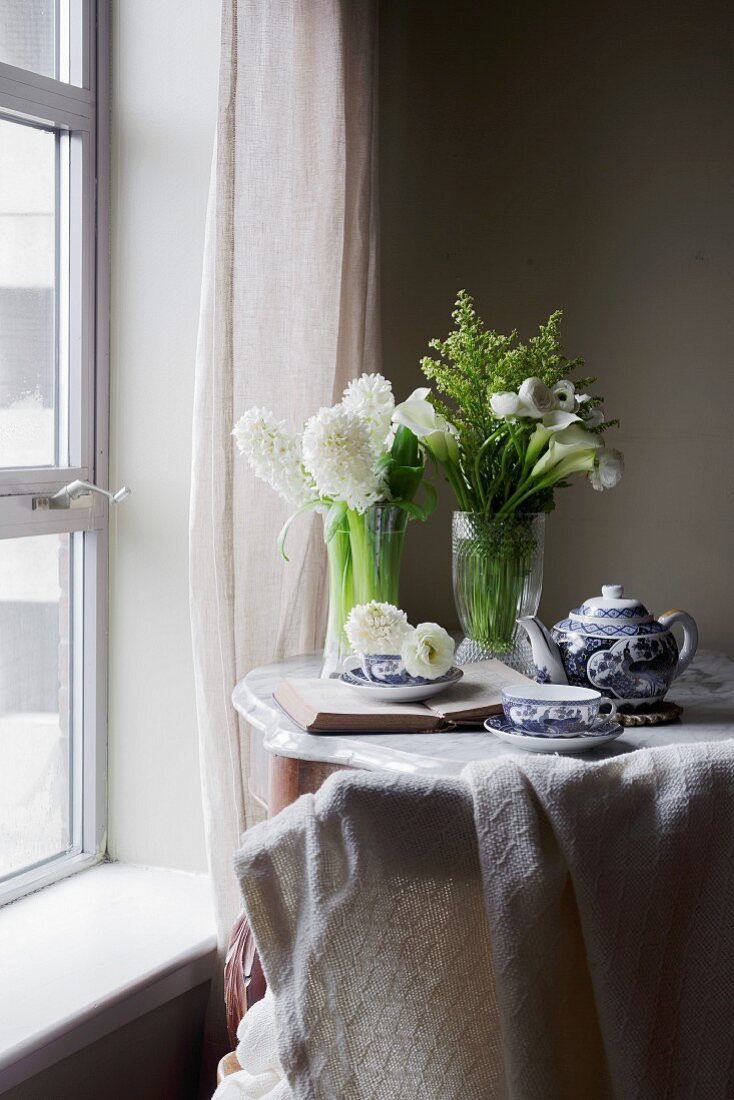 Frühstückstee mit Teetasse, Teekanne, weissen Blumen und Buch auf Tisch am Fenster