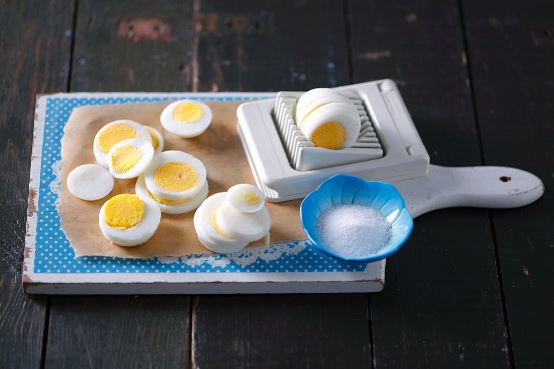 Sliced hard boiled eggs with an egg slicer and a bowl of salt on a wooden board