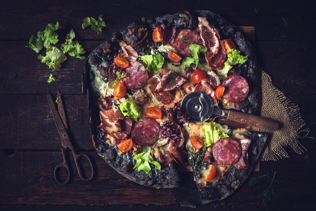 Black pizza with ham, sausage, bacon and vegetables on wooden background