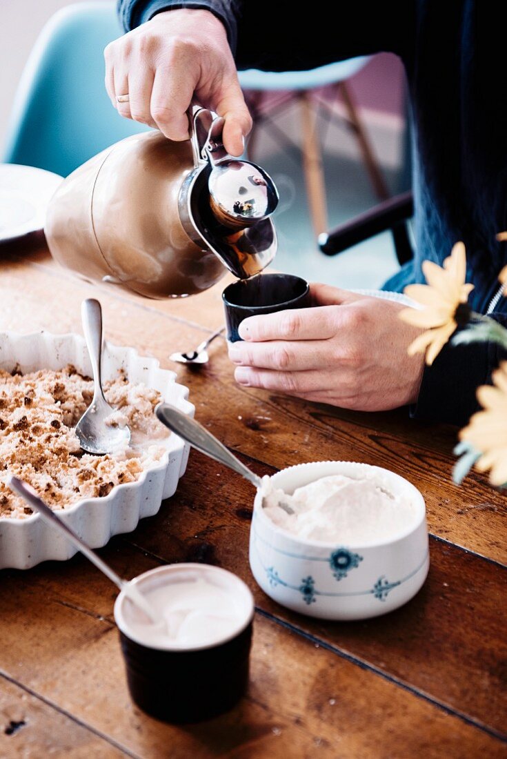 Apfelkuchen mit Sahne, im Hintergrund giesst Person Kaffee in Becher