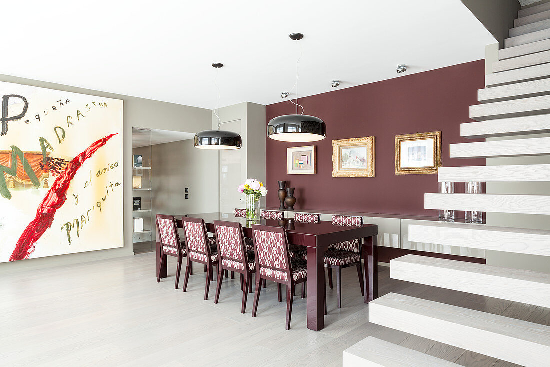 Upholstered chairs in elegant dining area with claret-red wall between huge artwork and cantilever staircase