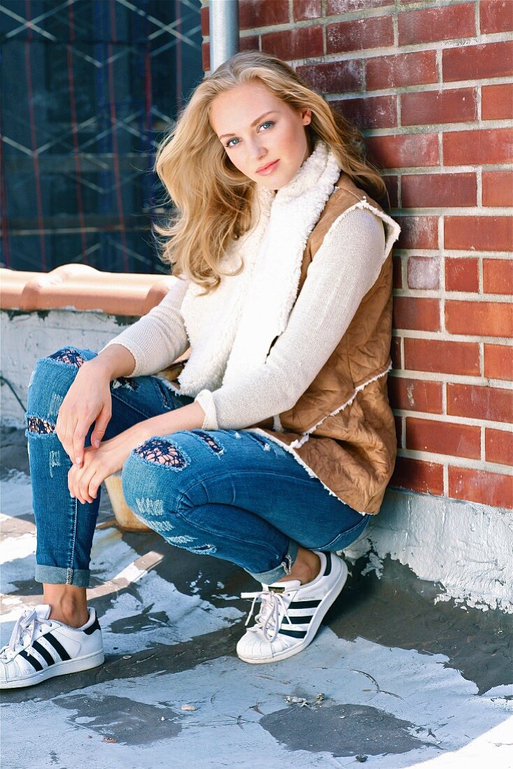 A blonde woman wearing a brown fur bodywarmer, a long-sleeved top, jeans and sneakers