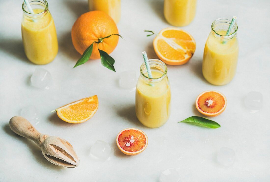 Healthy yellow smoothie with citrus fruit, ginger, ice in glass bottles over light marble table background