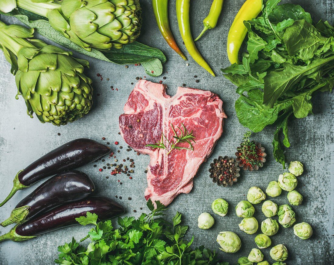 Rohes T-Bone-Steak umgeben von grünem Gemüse und Gewürzen (proteinreich)