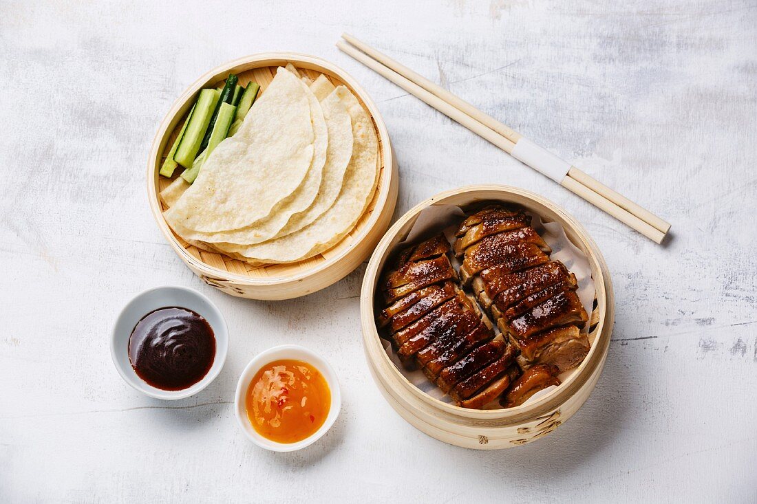 Peking Duck in bamboo steamer served with fresh cucumber, sauce and roasted wheaten chinese pancakes on white background