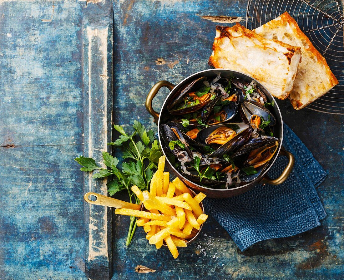 Mussels in copper cooking pan, baguette toasts and french fries on blue background