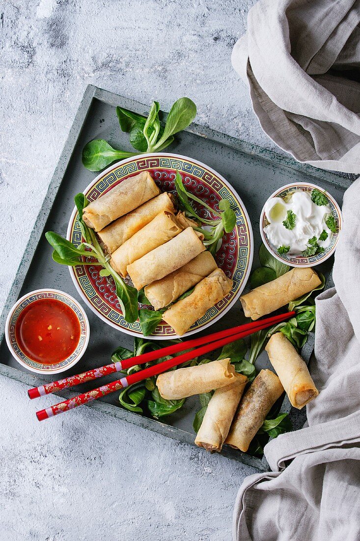 Fried spring rolls with red and white sauces, served in china plate on wood tray with fresh green salad and wooden chopsticks over gray blue texture background