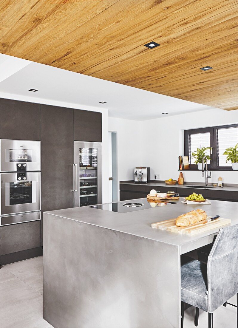 An anthracite-look island in front of a cupboard with built-in appliances and a suspended reclaimed wood ceiling