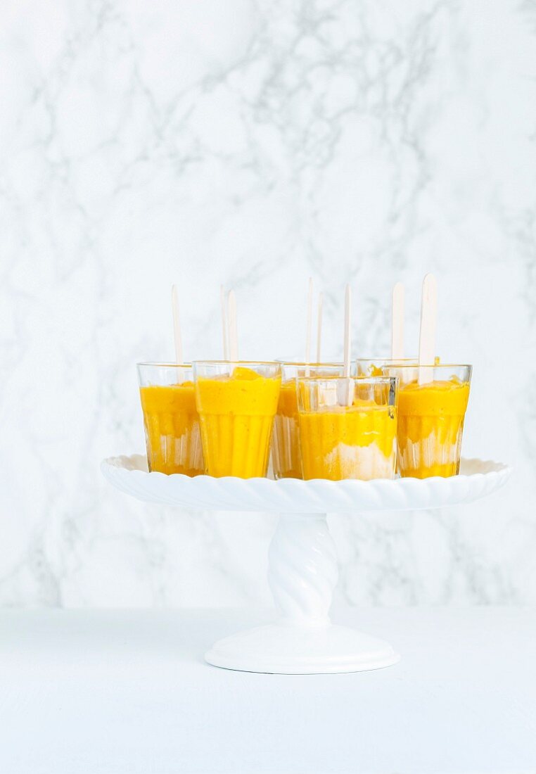 Mango-Popsicles in Gläsern auf Porzellanständer vor Marmorhintergrund