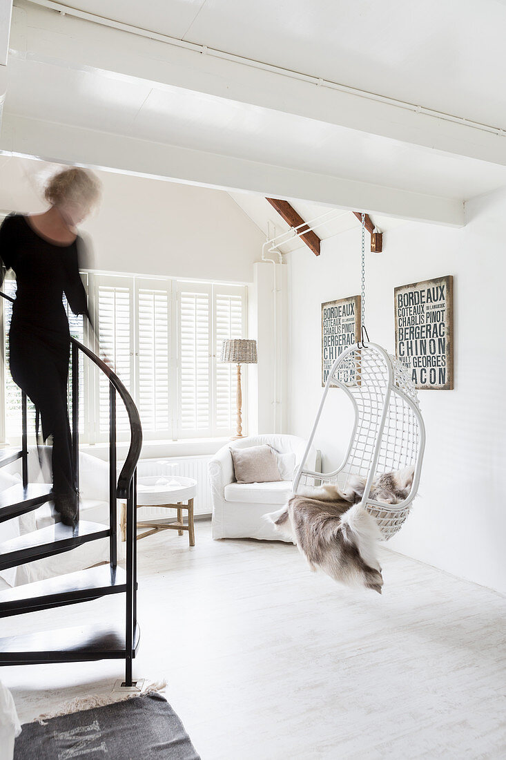 Woman walking down winding staircase into white living room with hanging chair