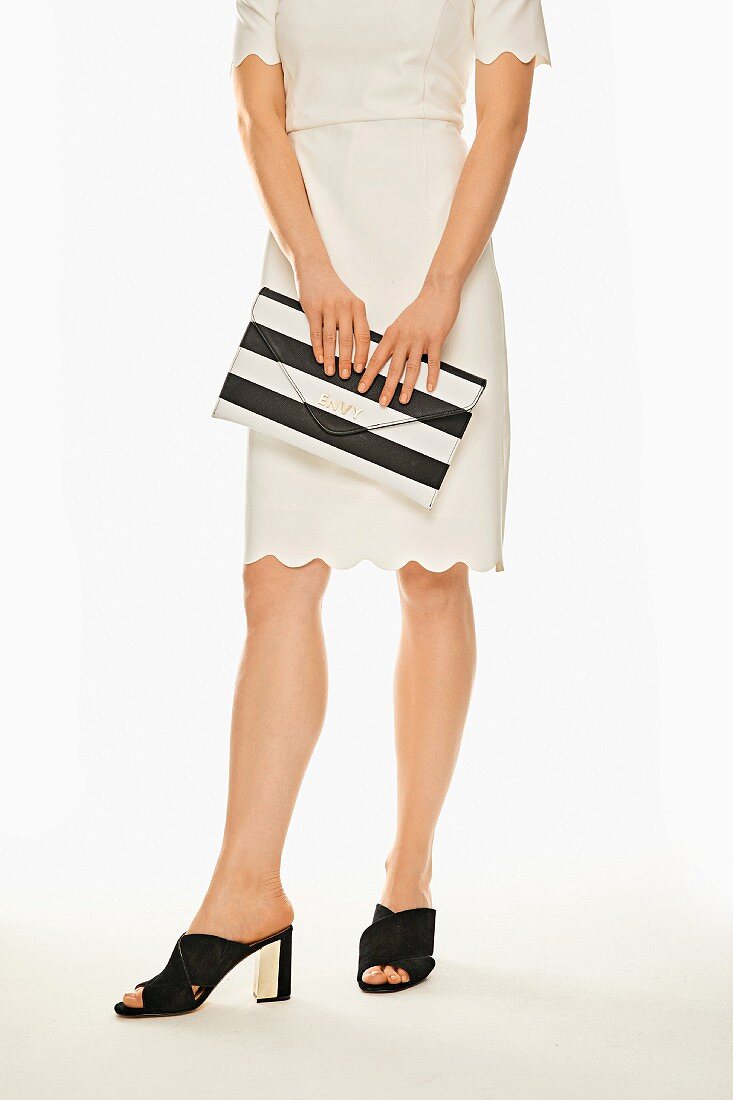 A woman wearing a white dress with a scalloped hem and sandals, carrying a black and white striped clutch
