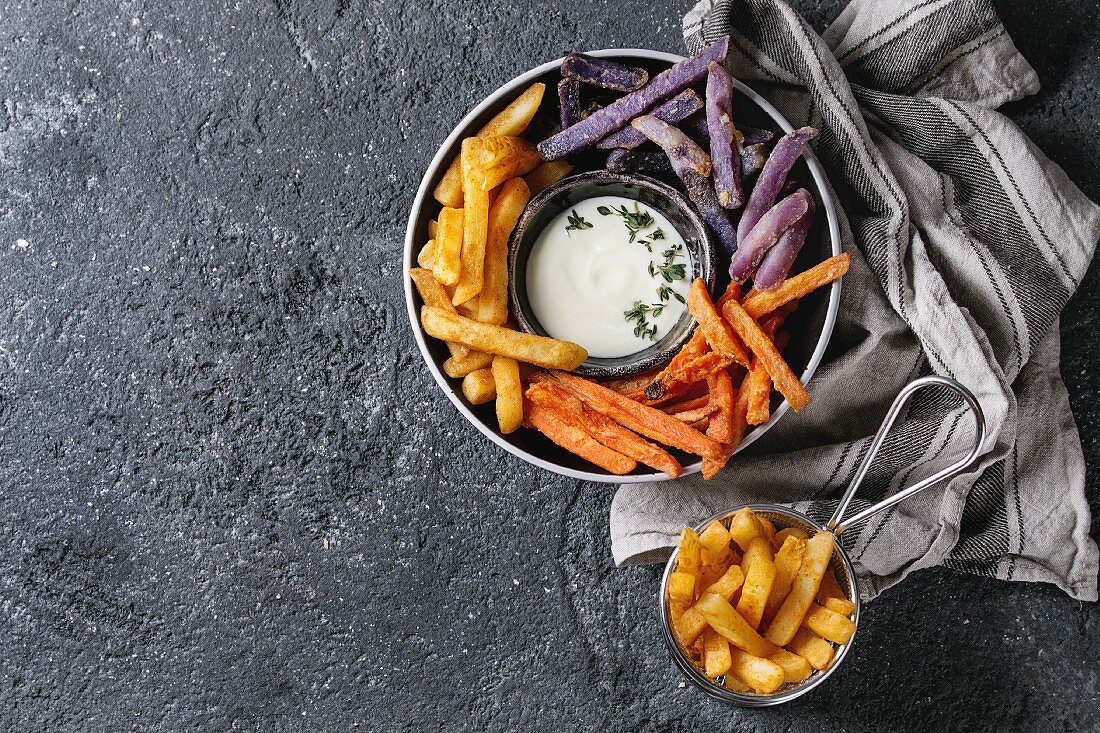 Variety of french fries traditional potatoes, purple potato, carrot served with white cheese sauce, salt, thyme in black bowl