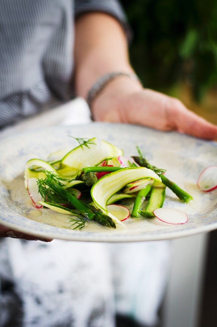 Hand hält einen Teller Zucchini-Spargel-Salat mit Radieschen und Dill