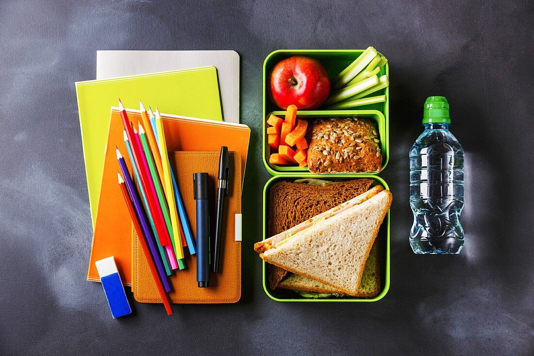 Sandwiches, Gemüse und Obst in Lunchbox, daneben Wasserflasche und Schulutensilien