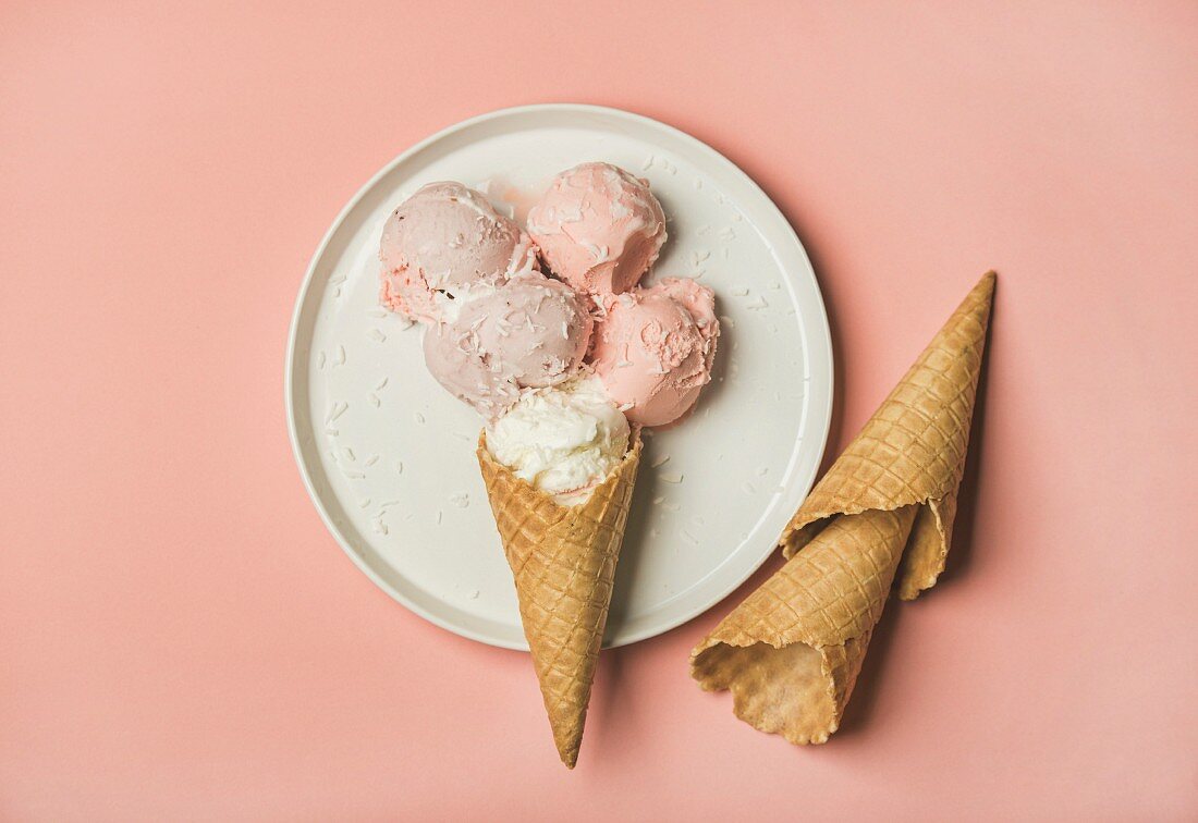 Flatlay of pastel pink strawberry and coconut ice cream scoops and sweet cones on white plate