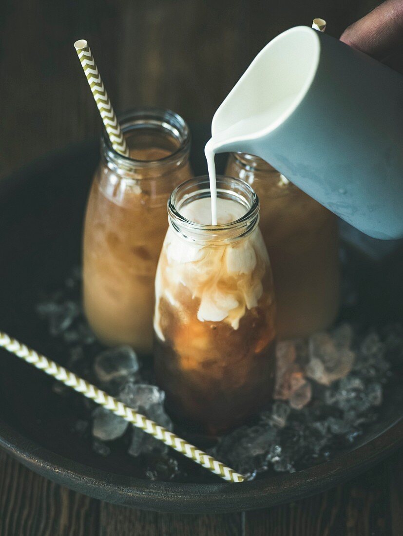 Cold Thai iced tea in glass bottles with milk pouring from jug on plate over dark wooden background