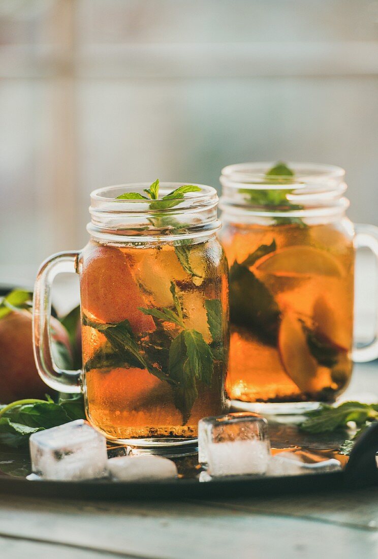 Summer refreshing cold peach ice tea with fresh mint leaves in glass jars on metal tray, selective focus