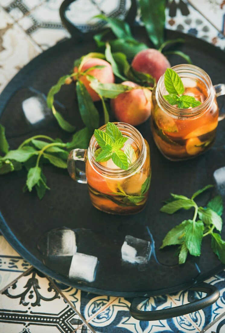 Summer refreshing cold peach ice tea with fresh mint in glass jars on metal tray over colorful oriental ceramic tile background