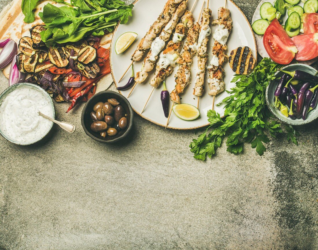 Flatlay of grilled chicken skewers with yogurt dip, flatbread, parsley, vegetables, marinated olives and chilis over grey concrete background