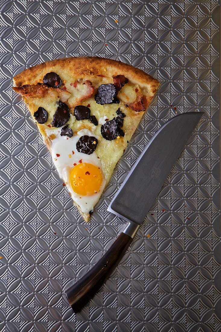 A slice of pizza with egg and black truffle on a stainless steel surface