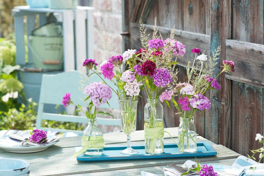 Tischdeko mit Dianthus ( Nelken ), Silene ( Roter Lichtnelke ), Lychnis coronaria ( Vexiernelke ) und Gräsern in Flaschen und Glas auf Holz-Untersetzer