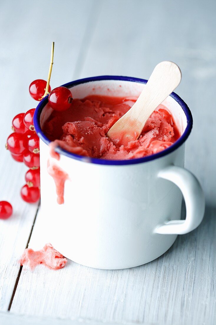 Red currant sorbet in an enamel cup with a wooden spoon and some fresh currants