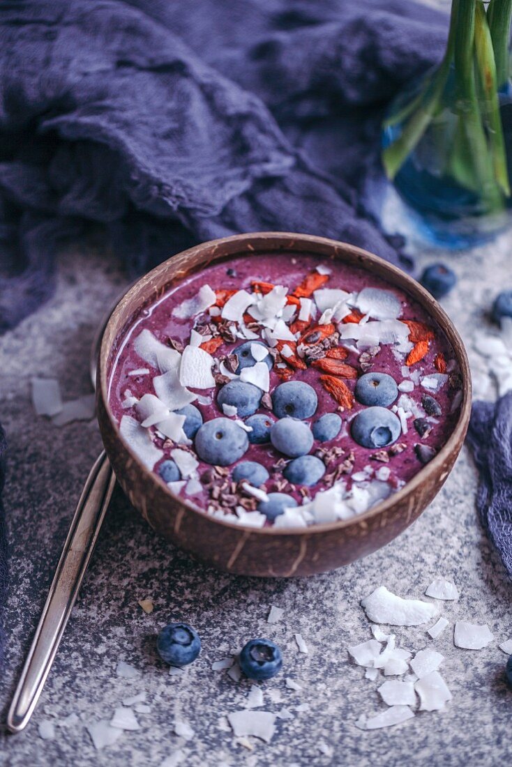 Blueberry smoothie topped with coconut flakes, goji berries, cacao nibs and blueberries in coconut bowl