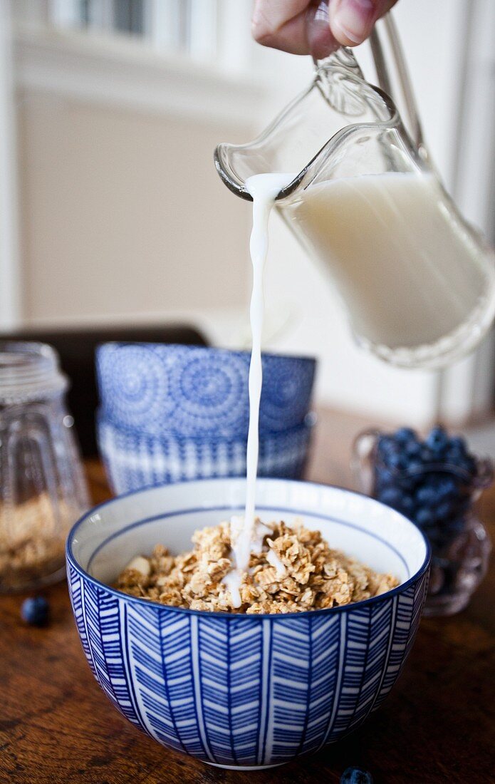 A woman pouring milk from a pitcher onto a bowl of granola, with blueberries, granola and a stack of bowls in the background