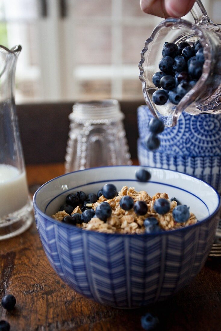 A woman pouring wild blueberries from a jar onto a bowl of granola with milk