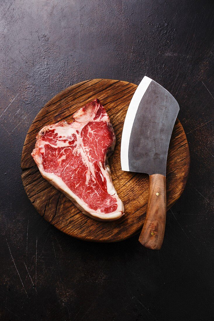 A raw dry-aged T-bone steak with a meat cleaver on a wooden board