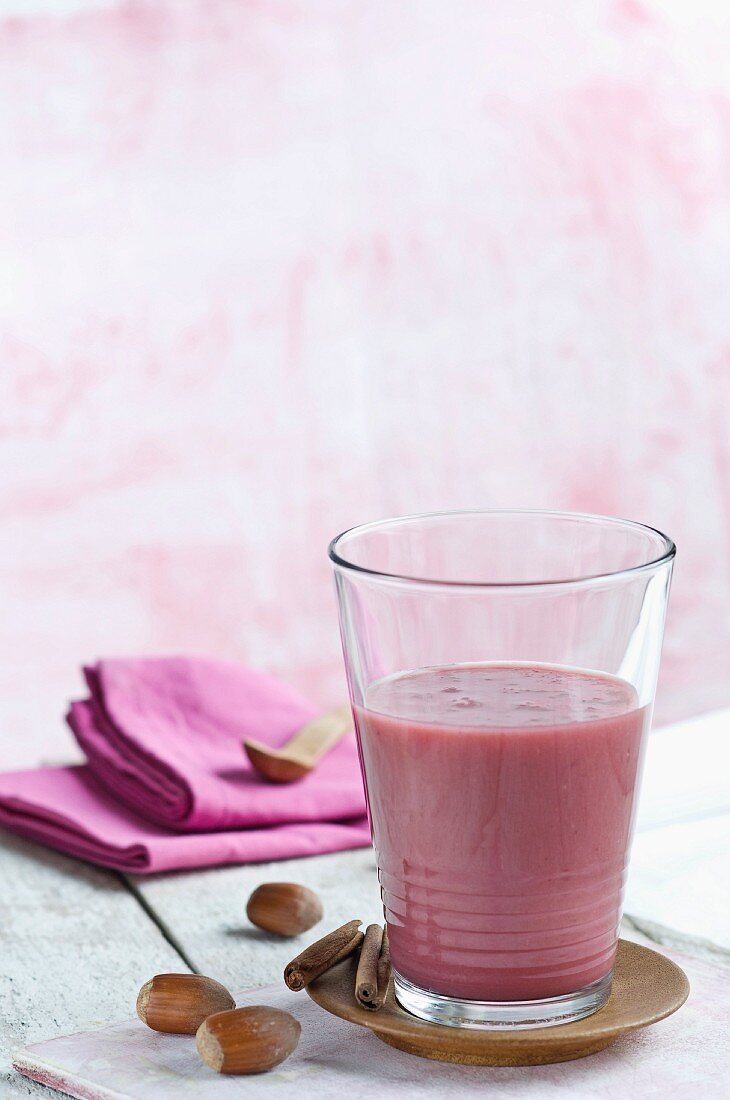 A berry smoothie in a glass next to hazelnuts and cinnamon sticks