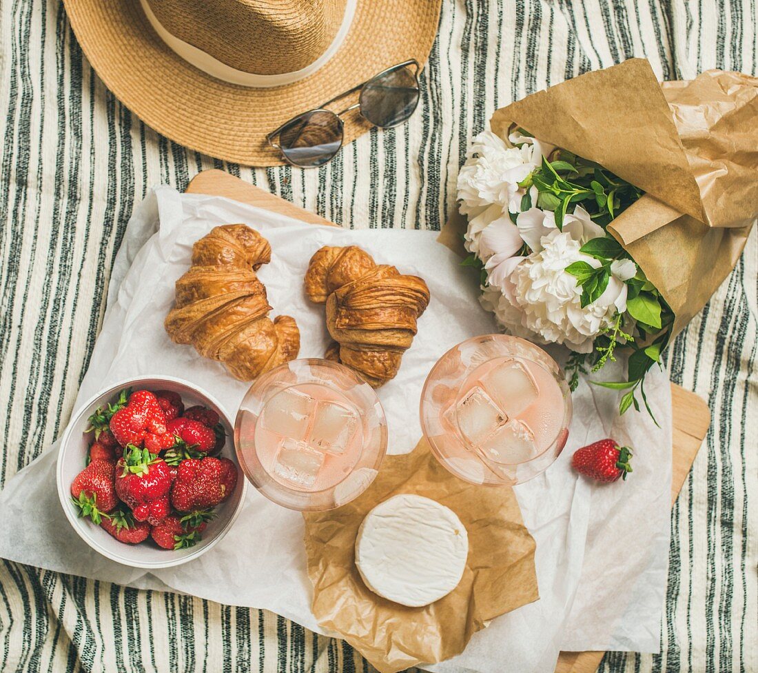 Rosé wine, strawberries, croissants, brie, a straw hat and a bouquet of flowers on a picnic blanket